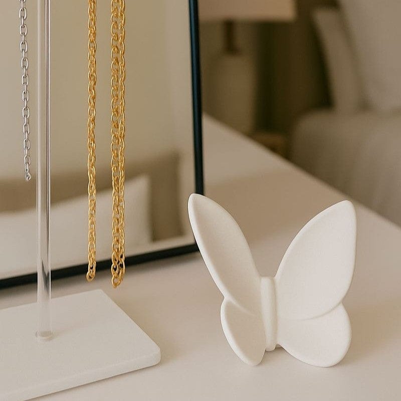 Matte white ceramic or resin butterfly sculpture/figurine displayed on a white bedside table next to a clear acrylic and metal jewelry stand holding gold and silver chain necklaces - Waterdale Collection