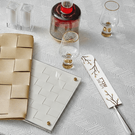 White embroidered leaf knife blade cover with a gold branch design and Hebrew inscription, resting on a geometric white tablecloth next to a challah knife - Waterdale Collection