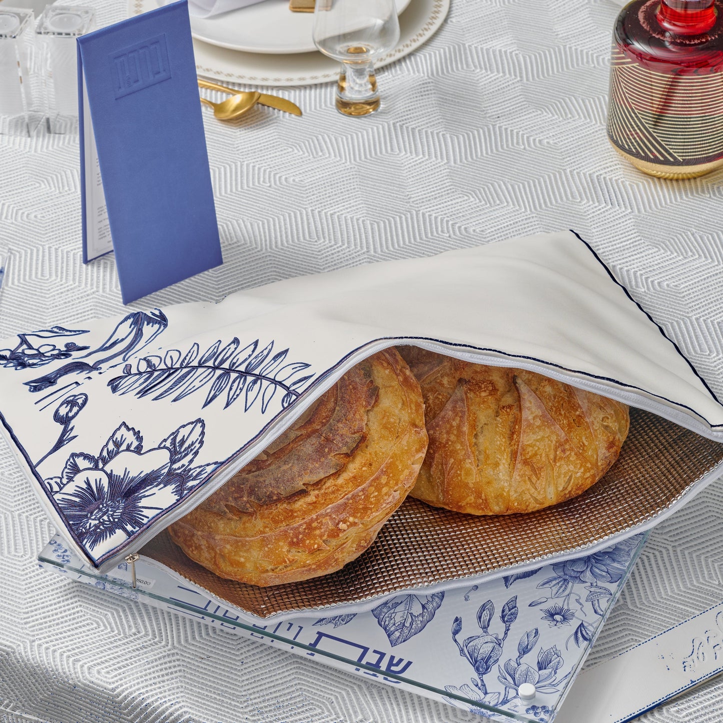 Blue French Toile Challah Pouch Cover styled on an elegant Shabbat table with gold-accented tableware and a blue bencher booklet.  