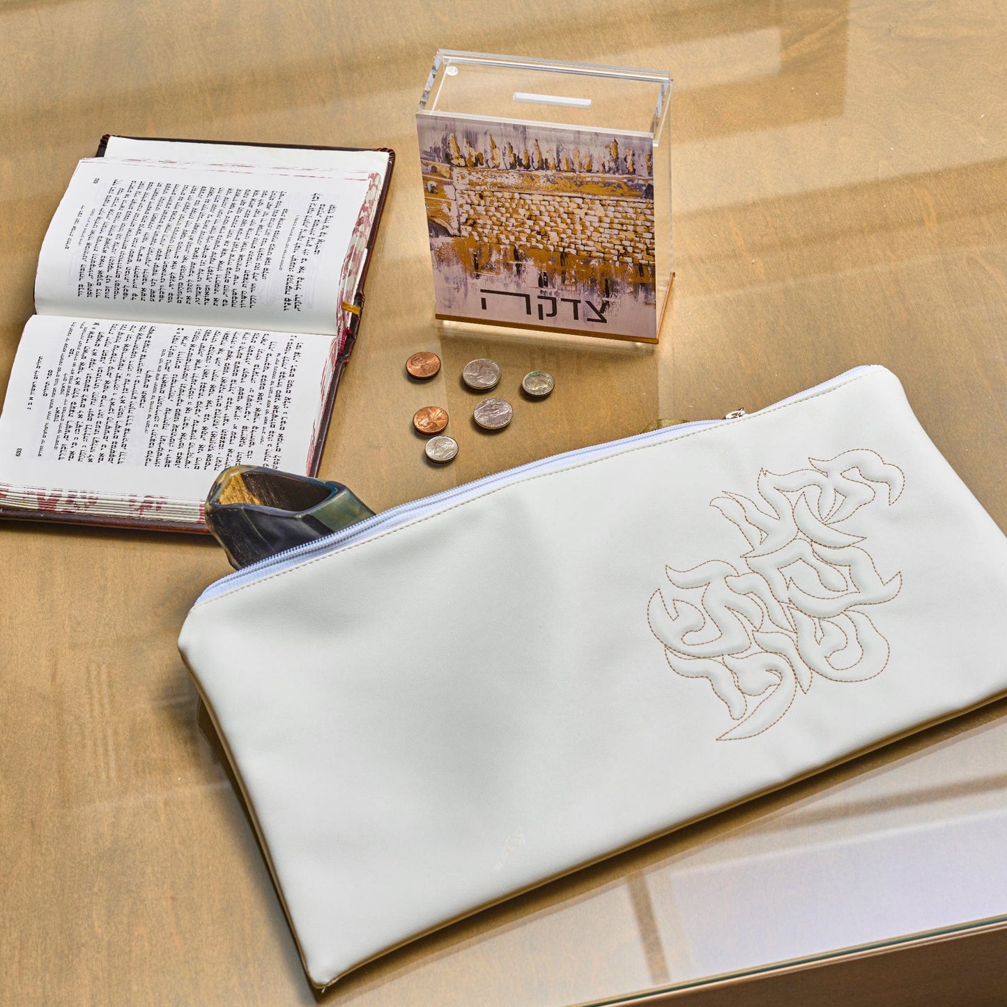 Kosel at Winter Tzedakah Box with white tefillin bag, open siddur prayer book, displayed on a glass table.