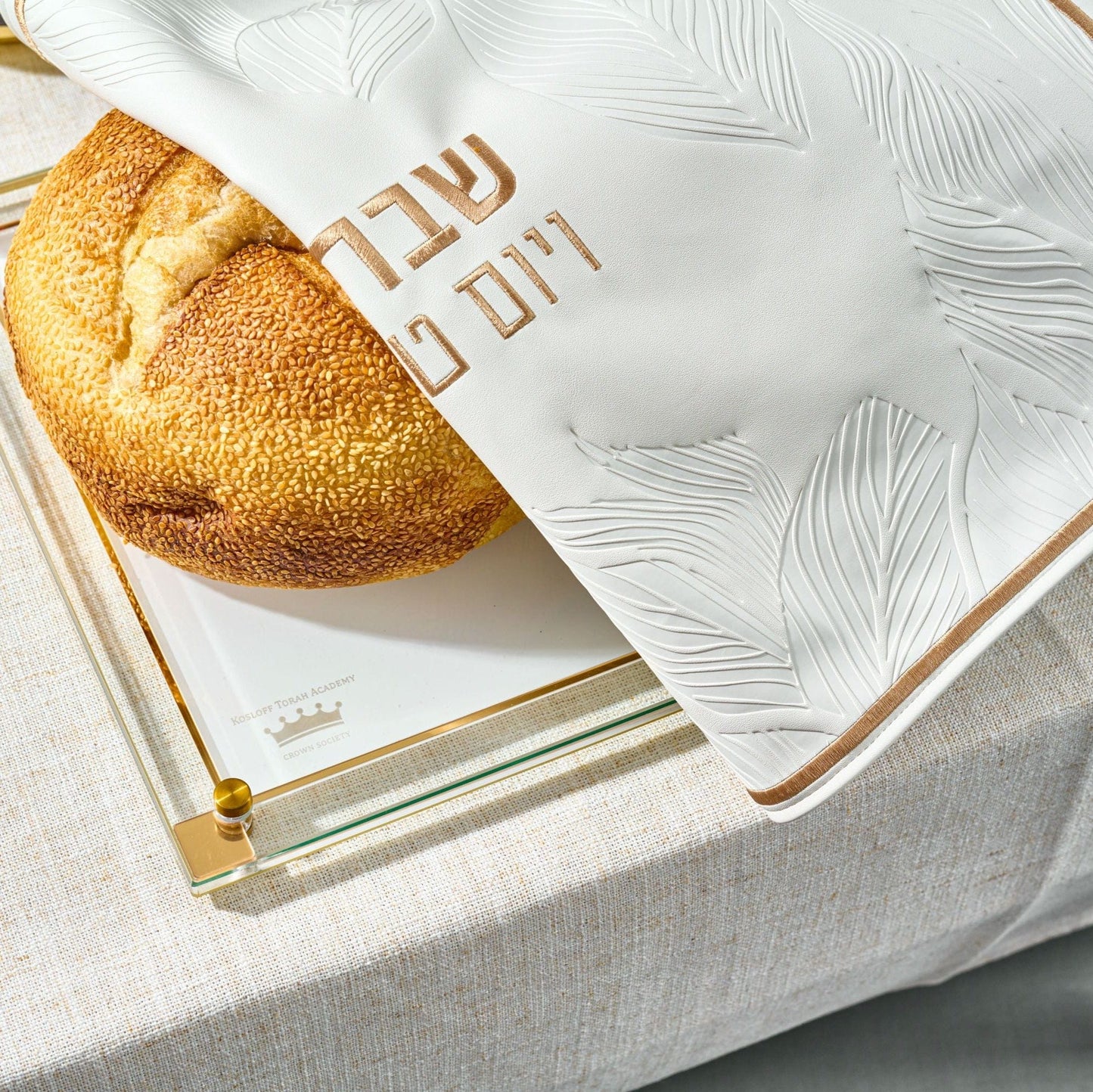 Round challah bread partially covered with Leaf Challah Cover on a glass challah board.