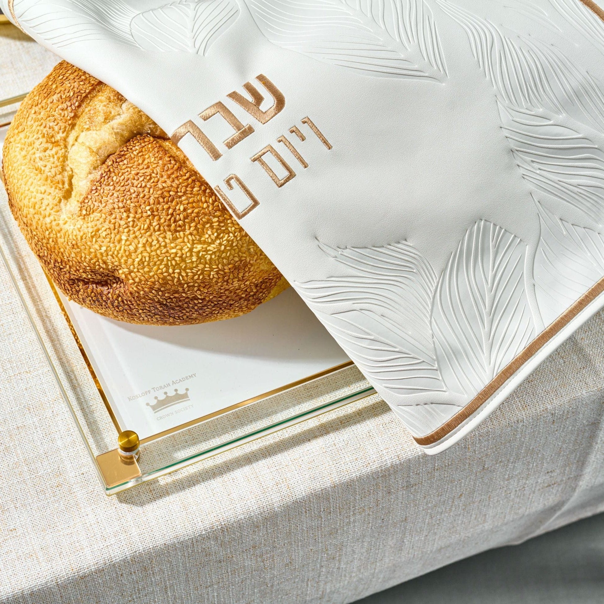 Round challah bread partially covered with Leaf Challah Cover on a glass challah board.