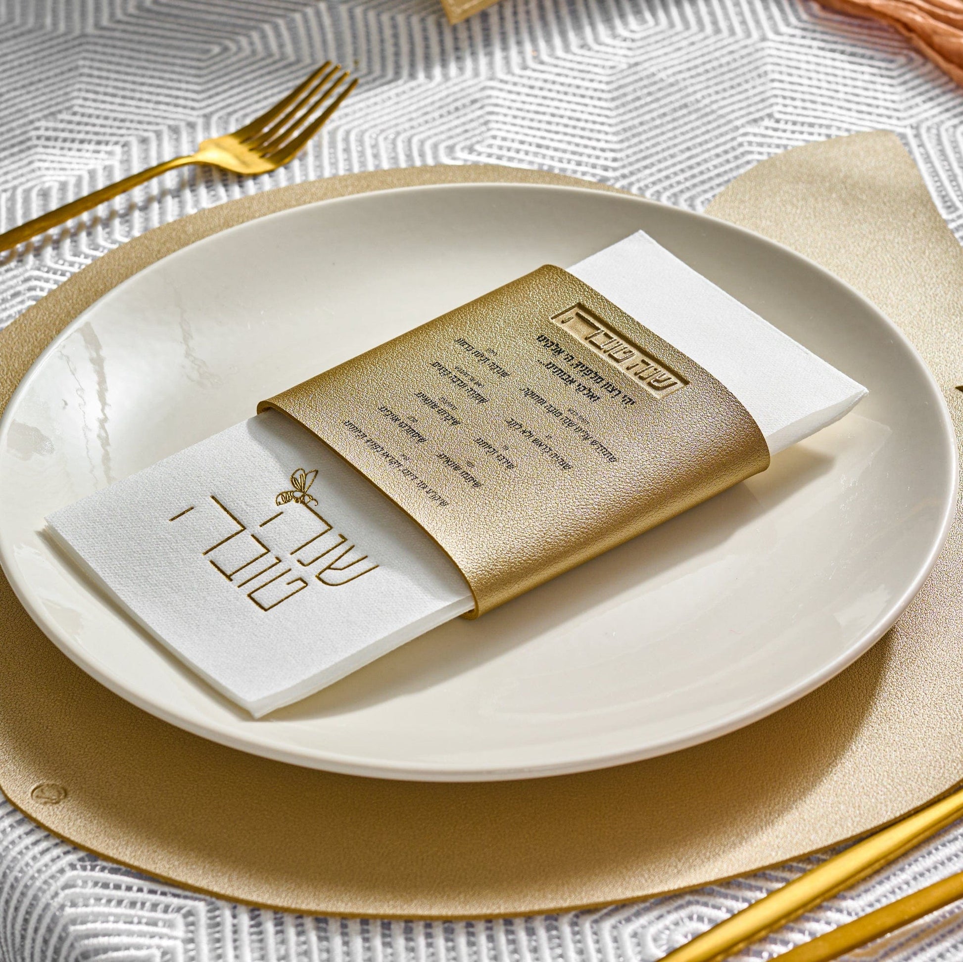 An elevated shot of a table setting featuring a Gold Leather Simanim Napkin Wrap on a white plate, leather gold apple charger and cutlery
