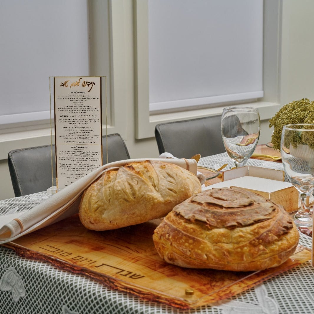 Wooden Challah Board on a Shabbat table setting with fresh challah loaves, folded challah cover, wine glasses, and a prayer card.